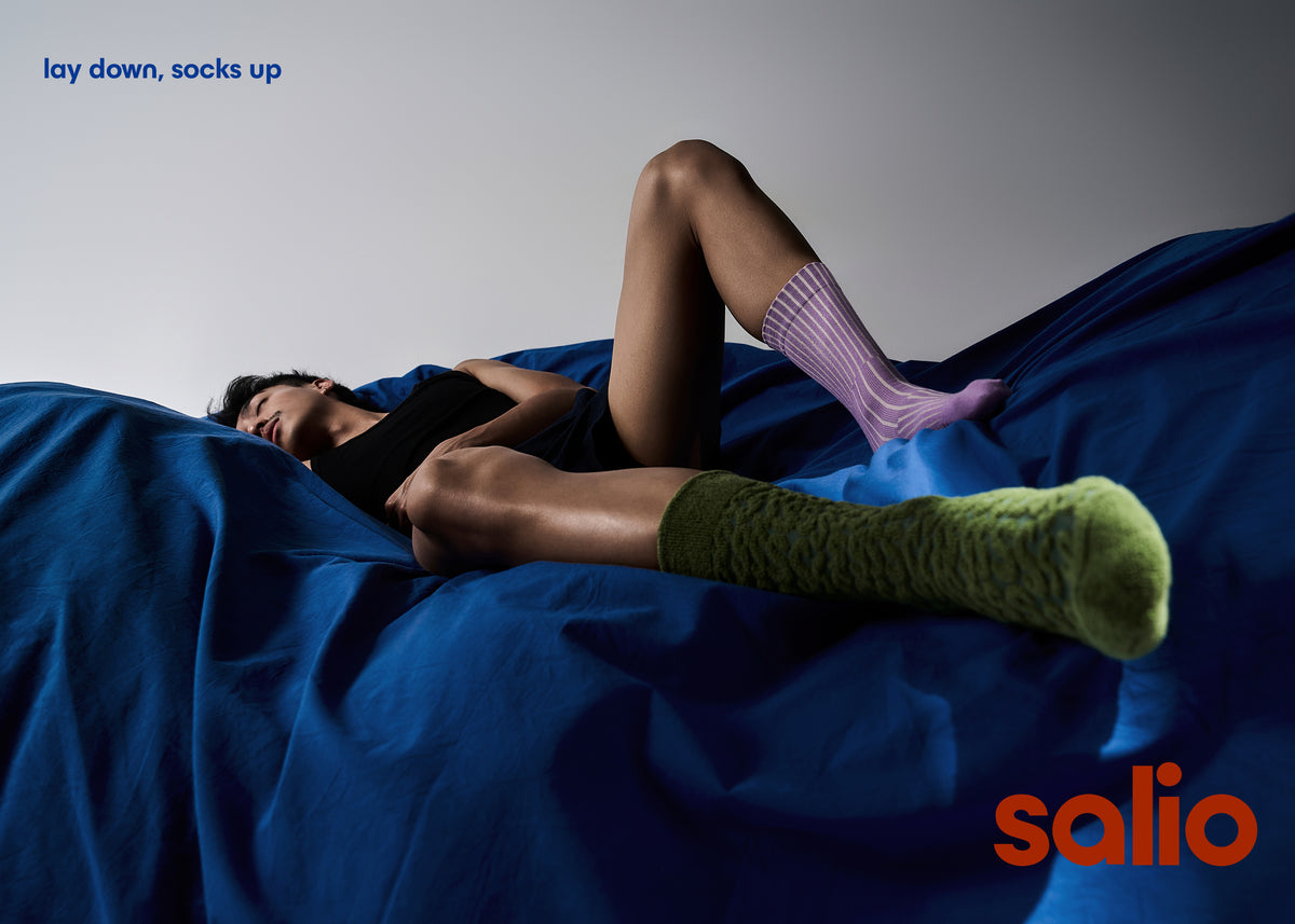 Model lying on a bed wearing the Coral Cotton Terry Socks in Color Forest on one foot and The Split Cotton Rib Socks in Color Lilac and Pale Grey on another foot. 