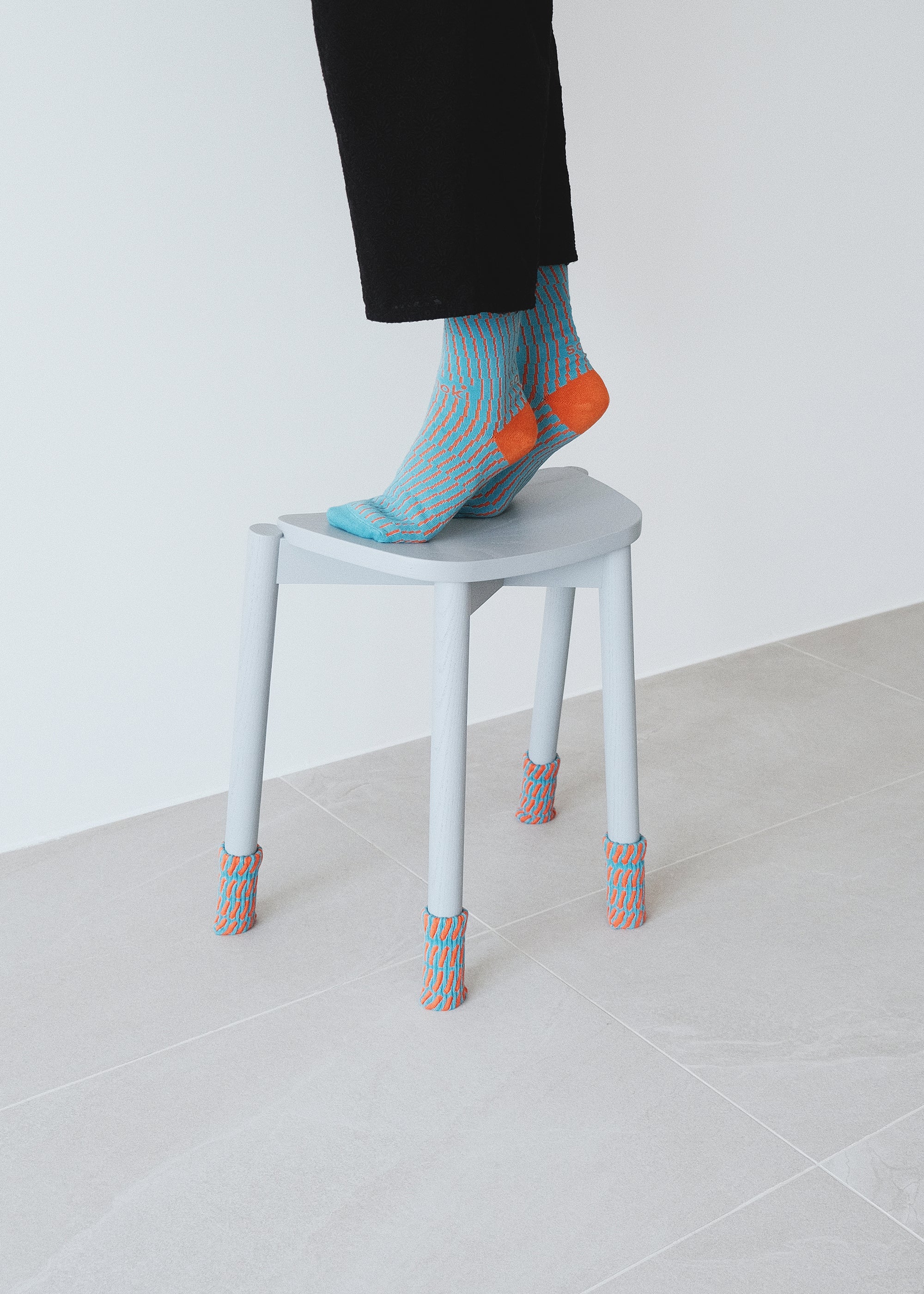 Person wearing The Swirling Gird Rib Socks in Color Cadet Blue and Tangeringe on a white stool with the Warp Chair Leg Cover in Color Cadet Blue and Tangerine